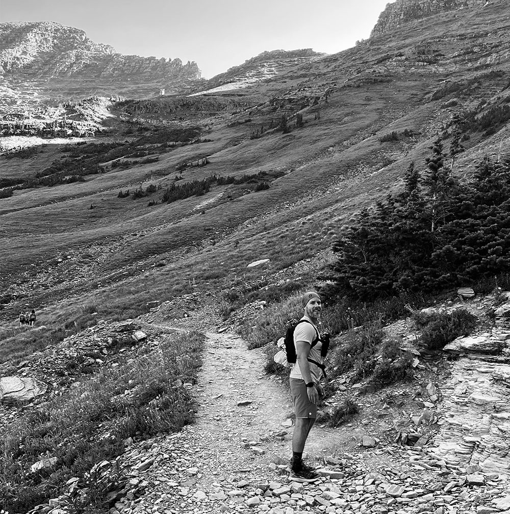 Black and white photo of a smiling man hiking a rocky mountain trail with a backpack, looking at the camera. Grassy slopes...