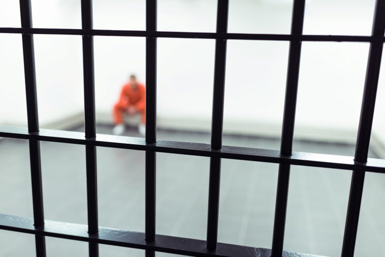 Prison bars in sharp focus in the foreground, with a blurred person in an orange jumpsuit sitting in a cell in the backgro...