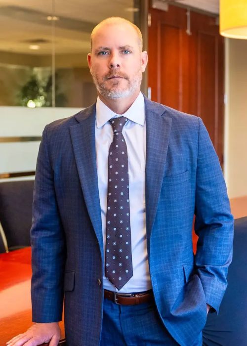 Portrait of a serious man in a blue suit with a patterned tie, standing in a corporate office setting with a conference ta...